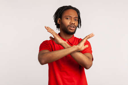 Portrait of young adult bearded man with dreadlocks wearing red T-shirt, crossing hands, gesturing warning or prohibition, meaning stop finish. Indoor studio shot isolated on gray background.の写真素材