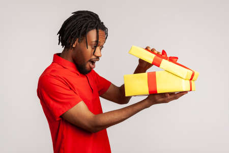 Amazed man with dreadlocks wearing red casual style T-shirt, opening gift box and peeping inside with excited facial expression, satisfied with present. Indoor studio shot isolated on gray background.の写真素材