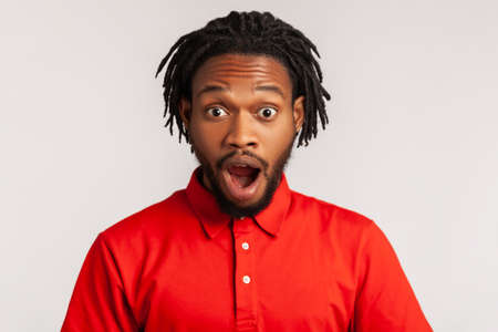 Astonished man with dreadlocks wearing red casual style T-shirt, standing with mouth open in surprise, has shocked expression, hears unbelievable news. Indoor studio shot isolated on gray background.の写真素材