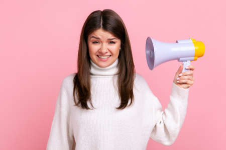 Portrait of young adult female holding megaphone, frowning face from loud sound, looking at camera, wearing white casual style sweater. Indoor studio shot isolated on pink background.の写真素材