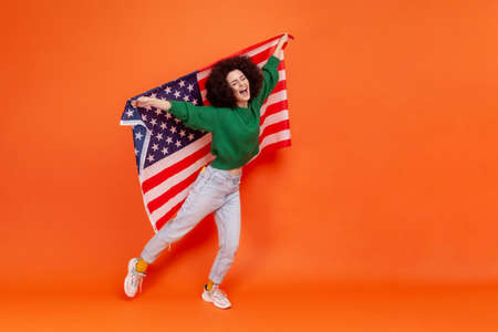 Full length photo of woman with Afro hairstyle wearing green casual style sweater raised arms, holding american flag, celebrating national holiday. Indoor studio shot isolated on orange background.の写真素材