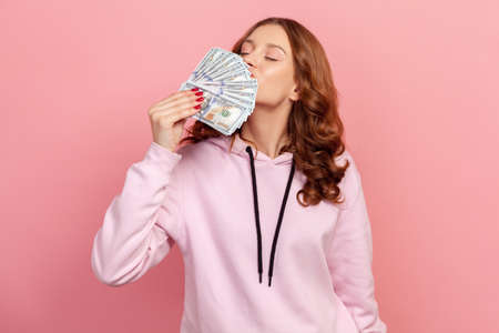 Portrait of pleased brunette teenage female sniffing dollar banknotes, enjoying lottery win, success. Indoor studio shot isolated on pink backgroundの写真素材