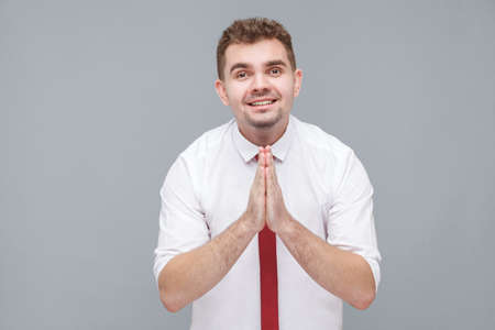 Please help me or forgive. Portrait of young hopeful man in white shirt and tie standing and looking at camera with pleased face pleading or asking for something. indoor isolated on gray background.の写真素材