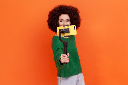 Young adult woman blogger with Afro hairstyle wearing green casual style sweater holding out steadicam with phone, making video or has livestream. Indoor studio shot isolated on orange background.の写真素材