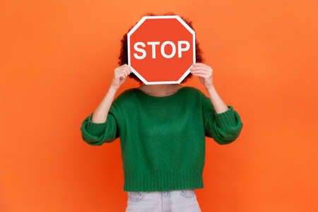 Unknown woman with wearing green casual style sweater hiding her face behind red stop traffic symbol, warning, avoids forbidden actions. Indoor studio shot isolated on orange background.の写真素材