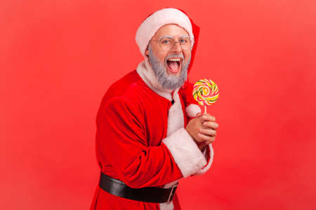 Portrait of happy amazed smiling elderly man with gray beard wearing santa claus costume standing with sugary lollypop in hands, looking at camera. Indoor studio shot isolated on red background.の写真素材
