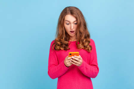 Portrait of amazed woman wearing pink pullover, looking at cellphone with surprised expression, reading shocking news using mobile phone. Indoor studio shot isolated on blue background.の写真素材