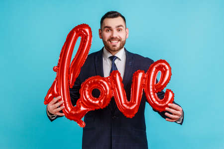 Portrait of happy smiling man wearing official style suit holding love word of foil balloons, expressing positive emotions and his feelings. Indoor studio shot isolated on blue background.の写真素材