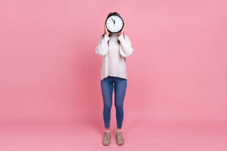 Full length portrait of unknown woman hiding her face behind big wall clock, showing time to go,...