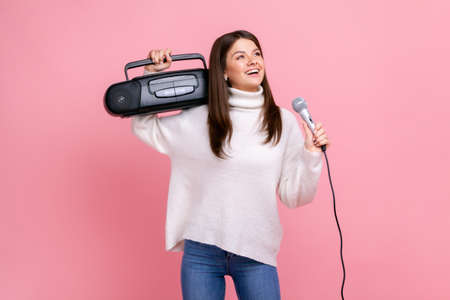 Portrait of happy positive female having fun, holding record player and singing via microphone, wearing white casual style sweater and jeans. Indoor studio shot isolated on pink background.の写真素材