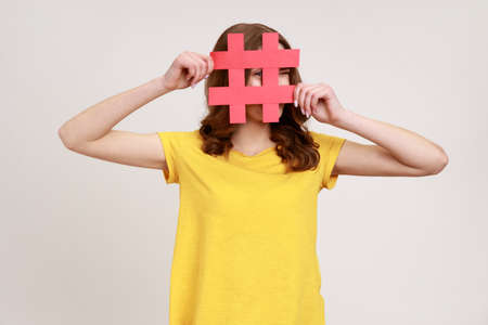 Curious brown haired woman of young age in yellow T-shirt looking through red hashtag sign, looking...