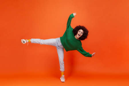 Full length portrait of amazed excited woman with Afro hairstyle in green casual sweater standing on one leg, having fun, raised arms and yelling. Indoor studio shot isolated on orange background.の写真素材