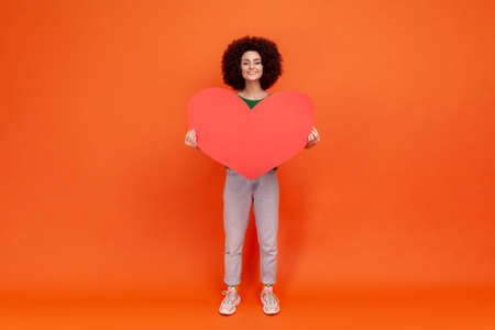 Full length portrait of falling in love woman with Afro hairstyle wearing green casual style sweater holding big red heart, looking with toothy smile. Indoor studio shot isolated on orange background.の写真素材