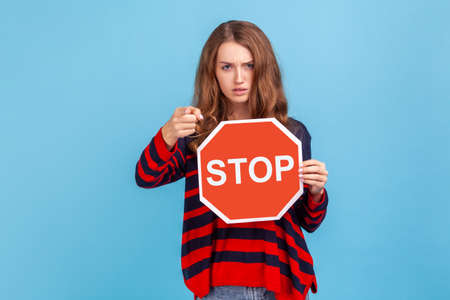 Serious woman wearing striped casual style sweater, holding big red stop road sign and pointing finger on you, strictly looking at camera. Indoor studio shot isolated on blue background.の写真素材