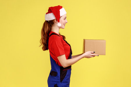 Side view portrait of courier female handing cardboard parcel for client, present for New Year, wearing blue overalls and santa claus hat. Indoor studio shot isolated on yellow background.の写真素材