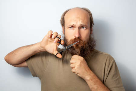 Adult handsome male cutting of his beard with scissors, looking away with regret in his eyes, bald...