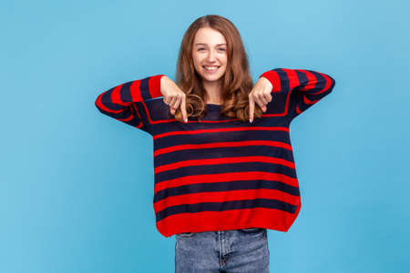 Look, advertise below. Cheerful woman wearing striped casual style sweater, pointing down place for commercial idea, looking at camera with toothy smile. Indoor studio shot isolated on blue backgroundの写真素材