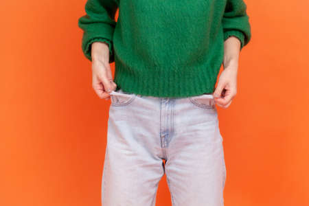 Unknown faceless woman wearing green casual style sweater showing empty pockets, has mo money, worried about debts and bankruptcy. Indoor studio shot isolated on orange background.の写真素材