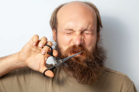 Portrait of male cutting of his beard, screws up his eyes, feels regret, holding scissors in hands,...