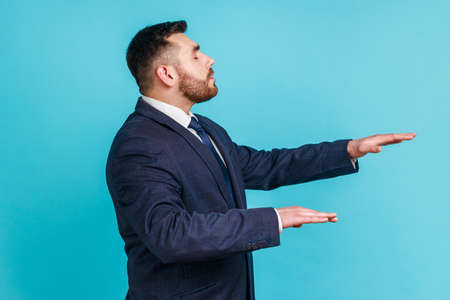 Side view of blind unhappy man wearing official style suit standing with closed eyes and touching air to find lost way, emulating walking in darkness. Indoor studio shot isolated on blue background.の写真素材