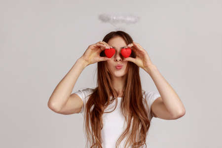 Woman covers eyes with toy hearts as if looking with love, sending air kiss, affection in relations, wearing white casual style T-shirt. Indoor studio shot isolated on gray background.の写真素材
