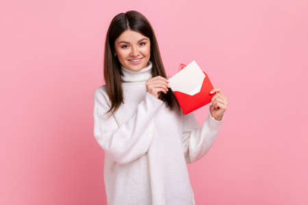 Portrait of woman standing, holding envelope and looking at camera with toothy smile and enjoying, wearing white casual style sweater. Indoor studio shot isolated on pink background.の写真素材