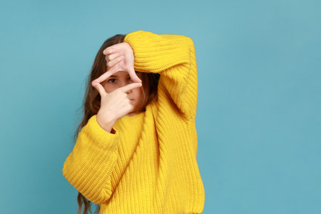 Portrait of little girl looking through photo frame hand gesture, focusing picture, observing world, wearing yellow casual style sweater. Indoor studio shot isolated on blue background.の写真素材