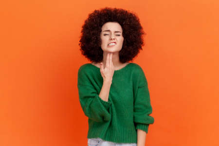 Portrait of woman with Afro hairstyle wearing green casual style sweater standing with finger gun, bored or tired, difficult to cope with problem. Indoor studio shot isolated on orange background.の写真素材