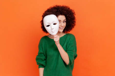 Woman with Afro hairstyle in green sweater removing white mask from face showing his smiling expression, good mood, pretending to be another person. Indoor studio shot isolated on orange background.の写真素材