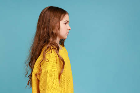 Side view portrait of serious smart bossy little girl with dark wavy hair looking straight ahead, wearing yellow casual style sweater. Indoor studio shot isolated on blue background.の写真素材