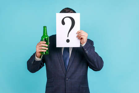 Portrait of anonymous person wearing dark official style suit hiding behind paper with question mark and holding bottle with alcohol. Indoor studio shot isolated on blue background.の写真素材