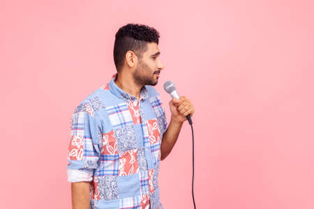 Side view of young adult handsome bearded man journalist standing holding microphone, wearing blue casual style shirt, looking ahead. Indoor studio shot isolated on pink background.の写真素材