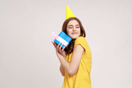 Portrait of happy satisfied teenager girl embracing blue gift box ,receiving surprise, being happy, keeps eyes closed, wearing yellow T-shirt. Indoor studio shot isolated on gray background.の写真素材
