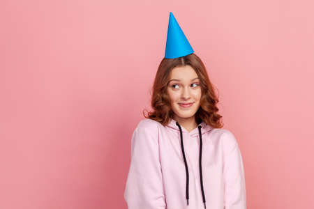 Portrait of shy curly haired teenage girl in hoodie and blue party hat looking up, birthday greetings. Indoor studio shot isolated on pink backgroundの写真素材