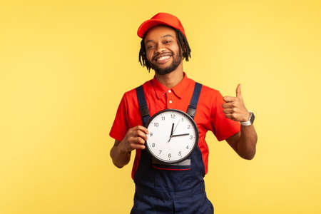 Happy punctual courier man in uniform holding clock and showing thumbs up, like gesture, satisfied with excellent delivery service on time. Indoor studio shot isolated on yellow background.の写真素材