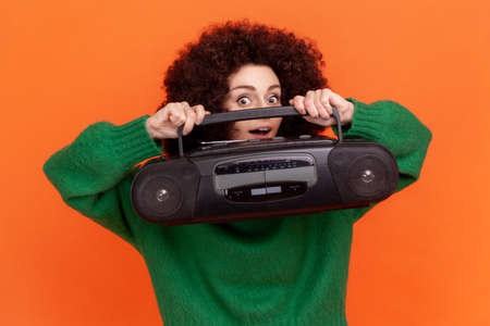 Happy funny woman with Afro hairstyle wearing green casual style sweater holding tape recorder in...