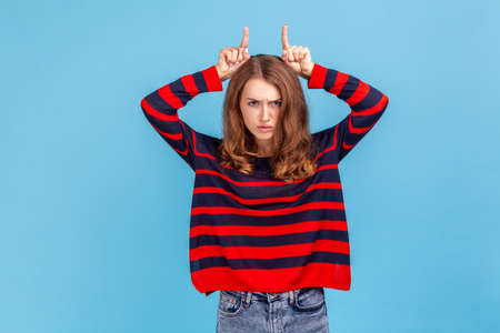 Woman showing bull horn gesture and looking with hate rage at camera, threatening and provoking conflict, wearing striped casual style sweater. Indoor studio shot isolated on blue background.の写真素材