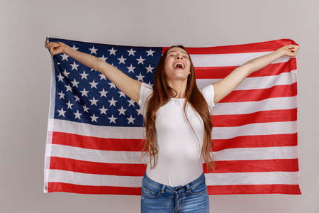 Portrait of attractive beautiful young woman with dark hair, holding USA flag over shoulders, rejoicing happily, wearing white T-shirt. Indoor studio shot isolated on gray background.の写真素材
