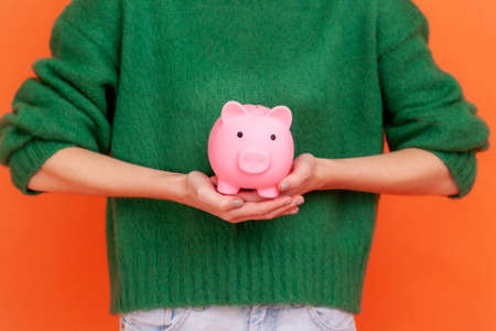 Unknown woman wearing green casual style sweater standing holding in hands piggy bank, investment,...