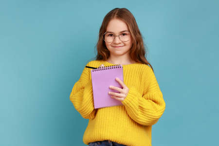 Portrait of positive little girl writing in notebook, looks smiling at camera, child doing homework, wearing yellow casual style sweater. Indoor studio shot isolated on blue background.の写真素材
