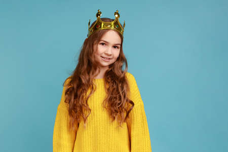 Portrait of cute little girl in golden diadem crown standing, looking at camera with toothy smile, wearing yellow casual style sweater. Indoor studio shot isolated on blue background.の写真素材