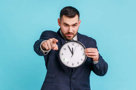Serious bearded man wearing official style suit pointing finger at camera holding big wall clock in hand, time to action, serious expression. Indoor studio shot isolated on blue background.の写真素材