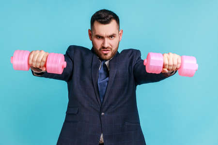 Strong bossy businessman wearing official style suit stretching arms with pink dumbbells, looking at camera with strict facial expression. Indoor studio shot isolated on blue background.の写真素材