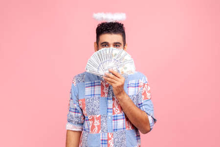 Angelic rich bearded man wearing blue casual style shirt with nimbus on head peeking out of dollar banknotes, rejoicing big profit earn. Indoor studio shot isolated on pink background.の写真素材