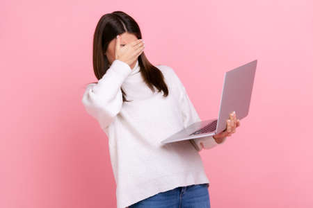 Portrait of brunette female holding laptop computer, covering eyes with palm, sees disgust content, wearing white casual style sweater. Indoor studio shot isolated on pink background.の写真素材