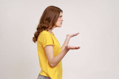 What do you want from me. Profile portrait of confused young female with brown hair in casual yellow T-shirt spreading hands, uncomfortable situation. Indoor studio shot isolated on gray background.の写真素材