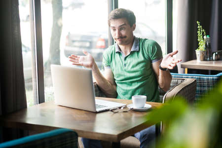 Portrait of confused man employee wearing green T-shirt, working online on laptop, shrugging...