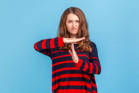 Portrait of stressed woman wearing striped casual style sweater showing time out gesture, looking with nervous expression, deadline. Indoor studio shot isolated on blue background.の写真素材