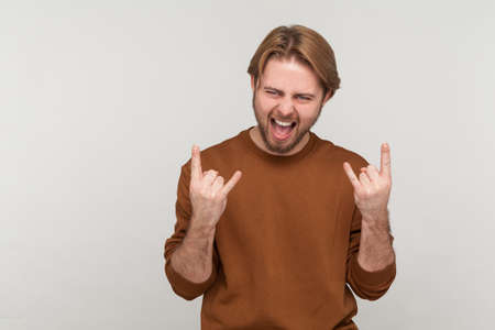Portrait of man with beard wearing sweatshirt, showing rock and roll sign hand gesture, screaming,...