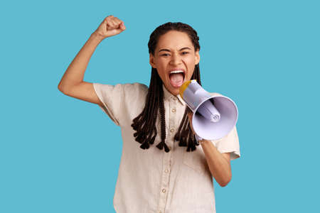 Portrait of woman with black dreadlocks standing with raised hands and holding megaphone, screaming...